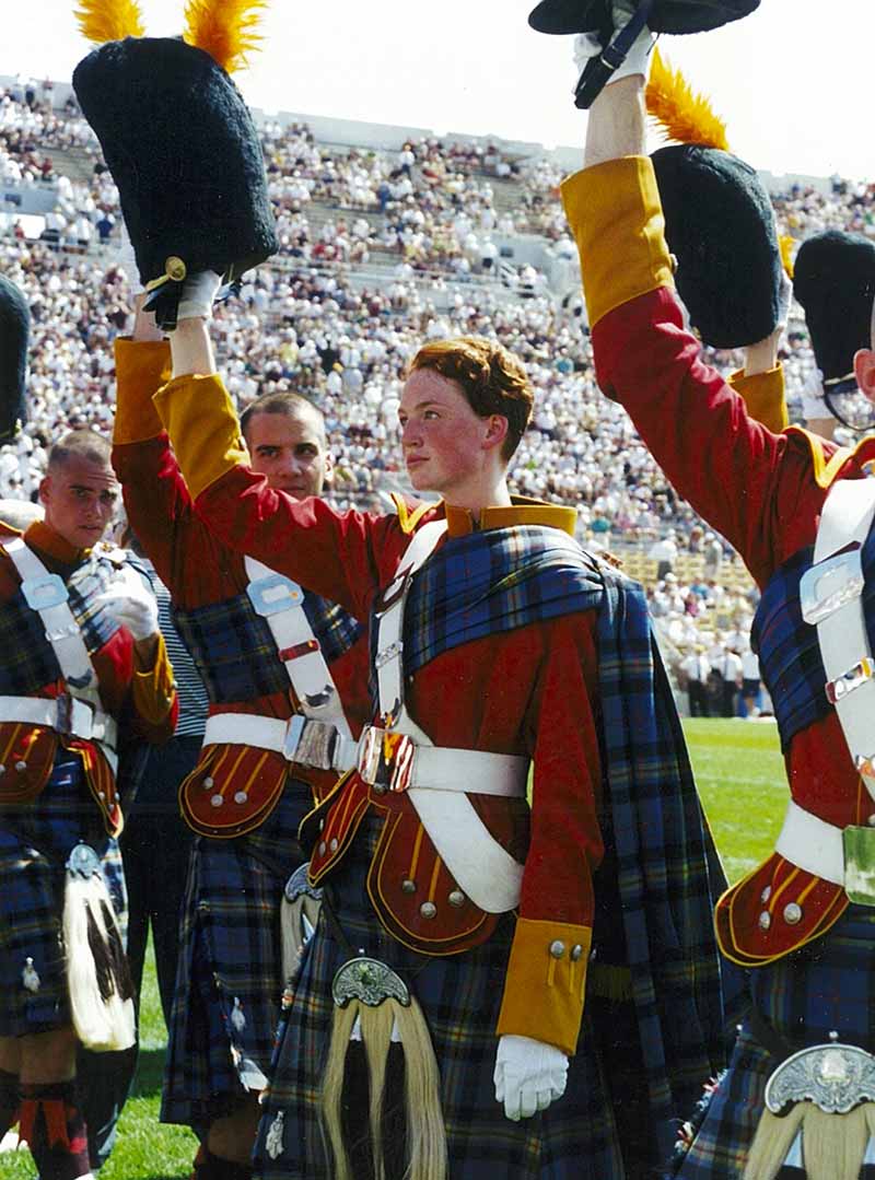 A woman raises her hat alongside the rest of the Irish Guard on the football field in their kilts.