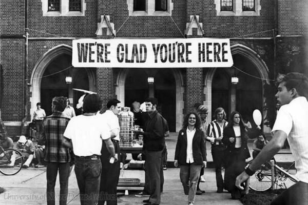 A group of students, including one woman looking towards the camera, stand in front of a large banner tacked onto a building that says "We're glad you're here".