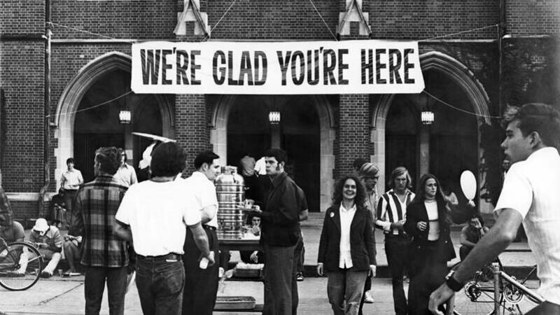 A group of students, including one woman looking towards the camera, stand in front of a large banner tacked onto a building that says 