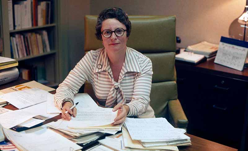 A woman with circle glasses works at a desk covered with papers.