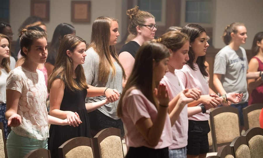 A room of female students hold hands and pray during a Mass.