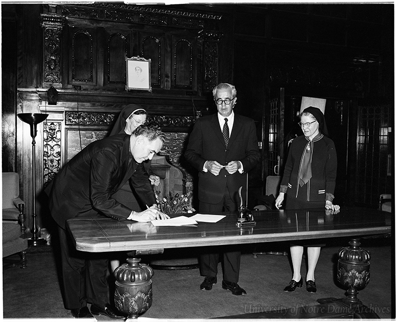 Fr. Hesburgh signs a document at a table, with 3 others.
