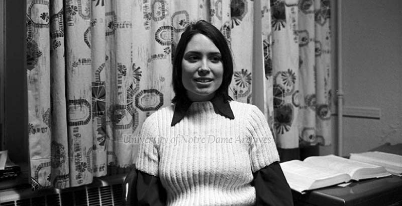 A black and white photo of a woman with dark hair in a white sweater in front of curtained window.
